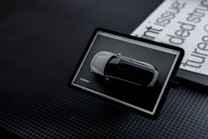 A tablet displays an image of a white car viewed from above. It is positioned on a dark, grid-patterned surface next to a stack of publications with black and white text partially visible.