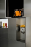 A modern-looking cabinet with a metallic finish. The cabinet has several compartments, one of which contains a brake disc with a yellow caliper. Another open compartment has a bag that is black with an orange front pocket. There are also three small items displayed in one of the compartments, which look like miniature models or packaged products.