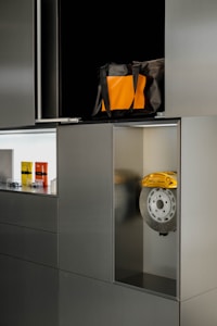 A modern-looking cabinet with a metallic finish. The cabinet has several compartments, one of which contains a brake disc with a yellow caliper. Another open compartment has a bag that is black with an orange front pocket. There are also three small items displayed in one of the compartments, which look like miniature models or packaged products.