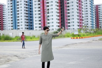 A person wearing sunglasses and a light green kurta stands on a road, posing with a camera. High-rise buildings with colorful facades, such as red, blue, and white, form the backdrop. Another person in a red shirt walks in the background, on a grassy area alongside the road.