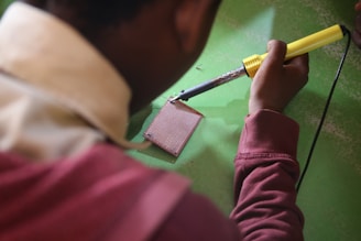 A person is using a soldering iron to work on a circuit board on a green surface. The person is wearing a maroon sweatshirt and the soldering iron has a yellow handle.