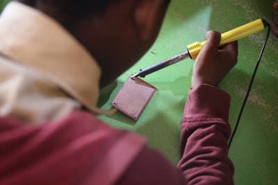 A person is using a soldering iron to work on a circuit board on a green surface. The person is wearing a maroon sweatshirt and the soldering iron has a yellow handle.