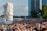 A large crowd of people is gathered near a waterfront where the iconic Merlion statue stands, spouting water. Several skyscrapers are visible in the background, and there are lush green trees offering some shade.