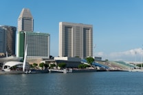 A waterfront cityscape featuring modern high-rise buildings, including a prominent hotel with a green facade. The foreground includes a body of water and a platform with sparse vegetation, and there appears to be an outdoor seating area nearby. The sky is clear and blue, contributing to a bright and sunny atmosphere.
