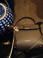 A taupe-colored Burberry handbag is placed on a wooden floor. It features the brand's logo embossed in gold. Above the handbag, there is a glimpse of a ceramic object with a blue and white geometric pattern.
