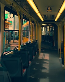 The interior of an empty public transportation vehicle is seen, likely a tram or bus, with yellow poles and overhead handrails. The seats are arranged in rows, and the windows allow natural light to create shadows on the floor. The view outside reveals urban surroundings with some signage visible.