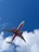 An airplane with a bright pink belly and wing accents flies overhead against a vibrant blue sky. The Brazilian flag is prominently displayed on the lower fuselage. Fluffy white clouds are scattered in the background, adding contrast to the scene.