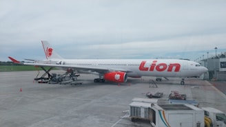 A large white and red commercial airliner is parked on the tarmac at an airport. The airplane is being serviced, with ground support equipment visible nearby, including a cargo loader. A few airport workers can be seen attending to the aircraft, and the background shows a grassy area and part of the airport terminal.