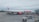 A large white and red commercial airliner is parked on the tarmac at an airport. The airplane is being serviced, with ground support equipment visible nearby, including a cargo loader. A few airport workers can be seen attending to the aircraft, and the background shows a grassy area and part of the airport terminal.