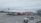 A large white and red commercial airliner is parked on the tarmac at an airport. The airplane is being serviced, with ground support equipment visible nearby, including a cargo loader. A few airport workers can be seen attending to the aircraft, and the background shows a grassy area and part of the airport terminal.