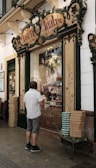 A man stands in front of a vintage-looking bakery shop with ornate signage. The display window shows various pastries and bread inside. The building exterior has decorative elements with floral motifs and green trim. Stacks of boxes are placed on a cart beside the man.