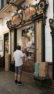 A man stands in front of a vintage-looking bakery shop with ornate signage. The display window shows various pastries and bread inside. The building exterior has decorative elements with floral motifs and green trim. Stacks of boxes are placed on a cart beside the man.