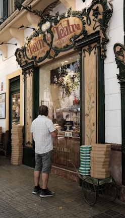 A man stands in front of a vintage-looking bakery shop with ornate signage. The display window shows various pastries and bread inside. The building exterior has decorative elements with floral motifs and green trim. Stacks of boxes are placed on a cart beside the man.