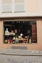 A shop window on a cobblestone street displays an eclectic assortment of items, including various pieces of furniture, ceramic pots, a lamp, and a decorative dog statue. The window frame and surrounding wall are painted a muted beige with bricks, and the upper section of the building features closed white shutters.