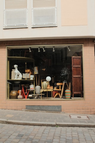 A shop window on a cobblestone street displays an eclectic assortment of items, including various pieces of furniture, ceramic pots, a lamp, and a decorative dog statue. The window frame and surrounding wall are painted a muted beige with bricks, and the upper section of the building features closed white shutters.