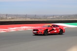 A red sports car with distinctive black and white patterns is speeding along a race track. The track is lined with barriers and fencing, and the background shows a cityscape under a bright, clear sky.
