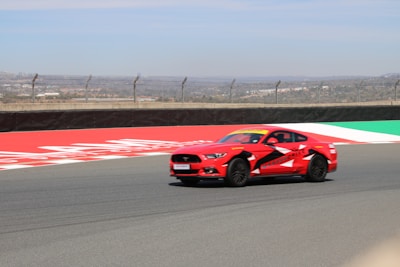A red sports car with distinctive black and white patterns is speeding along a race track. The track is lined with barriers and fencing, and the background shows a cityscape under a bright, clear sky.