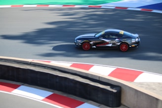 A sports car with a dark body and red wheels is racing on a track with red and white curbs. The track shows green and blue areas beyond the pavement. The car is branded with 'MASTER DRIVE' on its side.