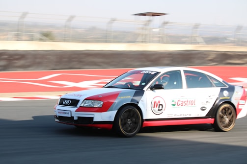 A racing car with sponsorship decals, including Castrol and MD, is speeding on a racetrack. It has a white and red color scheme, and the background shows part of the race track with a blurred motion effect.