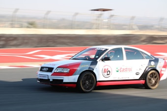 A racing car with sponsorship decals, including Castrol and MD, is speeding on a racetrack. It has a white and red color scheme, and the background shows part of the race track with a blurred motion effect.
