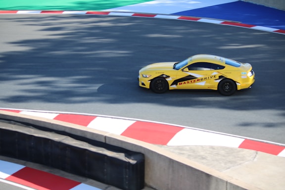 A yellow sports car with the marking 'MASTERDRIVE' on its side is navigating a racing track. The track is marked with red and white curbs, and there is a section of green and blue in the background. Shadows are cast onto the track surface, suggesting bright sunlight.