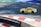 A yellow sports car with the marking 'MASTERDRIVE' on its side is navigating a racing track. The track is marked with red and white curbs, and there is a section of green and blue in the background. Shadows are cast onto the track surface, suggesting bright sunlight.