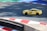 A yellow sports car with the marking 'MASTERDRIVE' on its side is navigating a racing track. The track is marked with red and white curbs, and there is a section of green and blue in the background. Shadows are cast onto the track surface, suggesting bright sunlight.