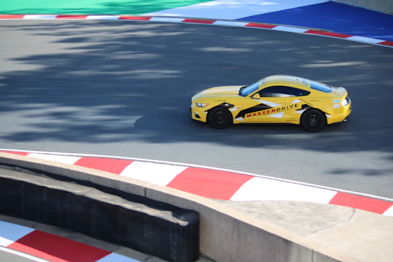 A yellow sports car with the marking 'MASTERDRIVE' on its side is navigating a racing track. The track is marked with red and white curbs, and there is a section of green and blue in the background. Shadows are cast onto the track surface, suggesting bright sunlight.