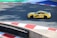 A yellow sports car with the marking 'MASTERDRIVE' on its side is navigating a racing track. The track is marked with red and white curbs, and there is a section of green and blue in the background. Shadows are cast onto the track surface, suggesting bright sunlight.