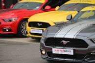 Three brightly colored sports cars are parked side by side. The cars are vibrant, featuring a red, a yellow, and a gray vehicle. Each car has the Mustang logo on the grille and a sporty appearance, with the 'MASTERDRIVE' sign attached to the front.
