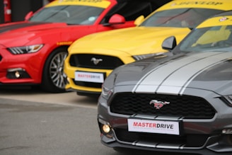 Three brightly colored sports cars are parked side by side. The cars are vibrant, featuring a red, a yellow, and a gray vehicle. Each car has the Mustang logo on the grille and a sporty appearance, with the 'MASTERDRIVE' sign attached to the front.