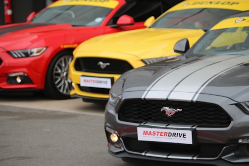 Three brightly colored sports cars are parked side by side. The cars are vibrant, featuring a red, a yellow, and a gray vehicle. Each car has the Mustang logo on the grille and a sporty appearance, with the 'MASTERDRIVE' sign attached to the front.