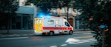 An emergency ambulance vehicle, marked 'PROTECTION CIVILE,' driving on a city street with flashing blue lights. The vehicle is white with red and yellow markings, and the surroundings include buildings and trees.