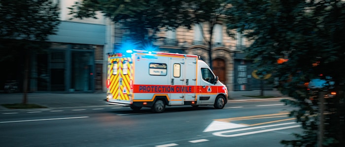 An emergency ambulance vehicle, marked 'PROTECTION CIVILE,' driving on a city street with flashing blue lights. The vehicle is white with red and yellow markings, and the surroundings include buildings and trees.