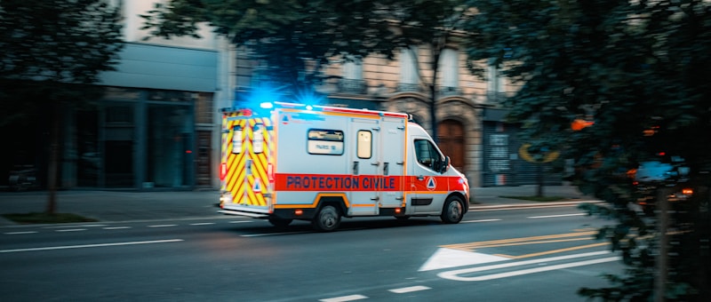 An emergency ambulance vehicle, marked 'PROTECTION CIVILE,' driving on a city street with flashing blue lights. The vehicle is white with red and yellow markings, and the surroundings include buildings and trees.