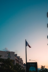 A French flag is prominently displayed on a flagpole against a backdrop of a clear sky transitioning from light blue to a warm hue at sunset. Below the flag, urban buildings with classic European architecture are visible, accompanied by lush green trees. The silhouette of a street sign with inscriptions is also present.
