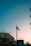 A French flag is prominently displayed on a flagpole against a backdrop of a clear sky transitioning from light blue to a warm hue at sunset. Below the flag, urban buildings with classic European architecture are visible, accompanied by lush green trees. The silhouette of a street sign with inscriptions is also present.