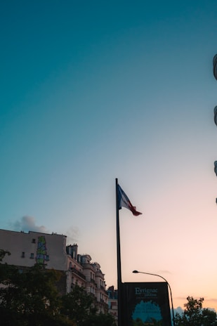 A French flag is prominently displayed on a flagpole against a backdrop of a clear sky transitioning from light blue to a warm hue at sunset. Below the flag, urban buildings with classic European architecture are visible, accompanied by lush green trees. The silhouette of a street sign with inscriptions is also present.