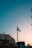 A French flag is prominently displayed on a flagpole against a backdrop of a clear sky transitioning from light blue to a warm hue at sunset. Below the flag, urban buildings with classic European architecture are visible, accompanied by lush green trees. The silhouette of a street sign with inscriptions is also present.