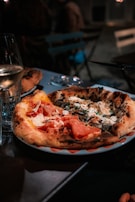 A plate of artisanal pizza sits on a table, featuring a variety of toppings including prosciutto, cheese, and herbs. Next to it, a glass of white wine stands, with blurrier details of a casual outdoor dining setup in the background.