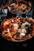 A close-up of a gourmet pizza with various toppings including prosciutto, cheese, herbs, and mushrooms. The pizza has a thick, golden-brown crust and is served on a colorful plate. In the background, there are wine glasses and water glasses, creating a dining atmosphere.