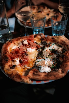 A close-up of a gourmet pizza with various toppings including prosciutto, cheese, herbs, and mushrooms. The pizza has a thick, golden-brown crust and is served on a colorful plate. In the background, there are wine glasses and water glasses, creating a dining atmosphere.