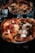 A close-up of a gourmet pizza with various toppings including prosciutto, cheese, herbs, and mushrooms. The pizza has a thick, golden-brown crust and is served on a colorful plate. In the background, there are wine glasses and water glasses, creating a dining atmosphere.