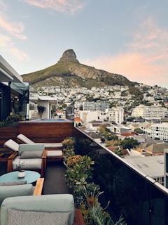A rooftop terrace overlooks a cityscape with a prominent, rugged mountain in the background. The terrace is elegantly furnished with cushioned wooden chairs and a small round table. Potted plants line the edge of the terrace, adding a touch of greenery to the urban setting. The buildings below are densely packed residential structures with a mix of modern and traditional designs.