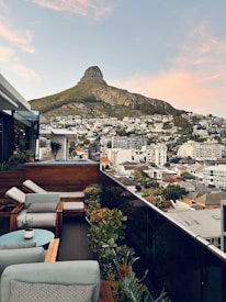 A rooftop terrace overlooks a cityscape with a prominent, rugged mountain in the background. The terrace is elegantly furnished with cushioned wooden chairs and a small round table. Potted plants line the edge of the terrace, adding a touch of greenery to the urban setting. The buildings below are densely packed residential structures with a mix of modern and traditional designs.
