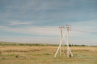 A rural landscape featuring several tall electricity poles with wires stretching horizontally across a grassland. The land appears dry with patches of grass and a few distant trees under a clear blue sky.