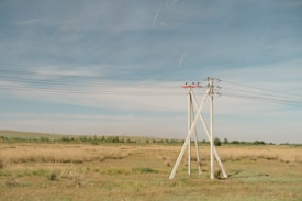 A rural landscape featuring several tall electricity poles with wires stretching horizontally across a grassland. The land appears dry with patches of grass and a few distant trees under a clear blue sky.