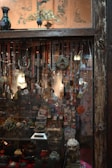 A richly decorated shop window displays an array of hanging necklaces and beads, with intricate ceramic and wooden decorations interspersed. Below, a variety of small antique objects, including vases, figurines, and ornate containers, fill the shelf. A colorful peacock sculpture adorns the top of the frame, adding a touch of elegance and color against the muted background, which is decorated with faded wall art. The overall scene is dimly lit, enhancing the rustic and vintage feel.