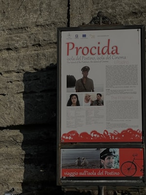 A framed poster is mounted on a textured stone wall. The poster features the word 'Procida' prominently in red, with information about 'Il Postino', including images from the film and a depiction of a bicycle.