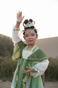 A child dressed in traditional attire with intricate patterns and green hues stands in an outdoor setting. The hair is styled in an elaborate updo with decorative ornaments. The background features natural elements like plants and shrubs, and the lighting suggests a warm, ambient atmosphere.